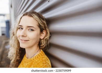 Young blonde girl standing in front of metal wall looking at camera smiling