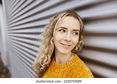 Young blonde girl stands in front of metal wall and looks to the side smiling