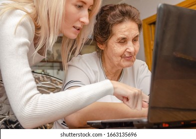 Young Blonde Girl Teaches An Elderly Dark-haired Woman, Grandmother How To Work On A Laptop At Home