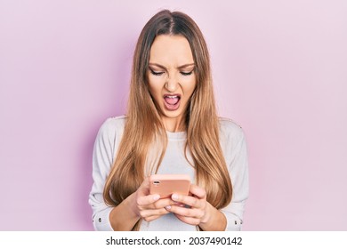 Young Blonde Girl Using Smartphone Typing Message Angry And Mad Screaming Frustrated And Furious, Shouting With Anger. Rage And Aggressive Concept. 