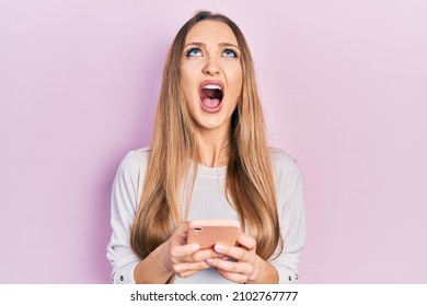 Young Blonde Girl Using Smartphone Typing Message Angry And Mad Screaming Frustrated And Furious, Shouting With Anger Looking Up. 