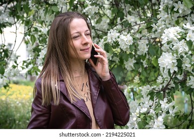 A Young Blonde Girl Walking In The Park Speaks On A Smartphone, The Spring Season Is The Blossoming Of The Apple Tree, People Use Technology.