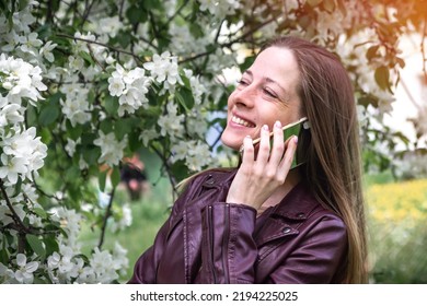 A Young Blonde Girl Walking In The Park Speaks On A Smartphone And Laughs, The Spring Season Is The Blossoming Of The Apple Tree, People Use Technology.