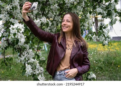 A Young Blonde Girl Walking In The Park Makes A Selfie On A Smartphone And Laughs, The Spring Season Is The Blossoming Of The Apple Tree, People Use Technology.