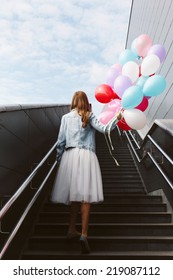 Young Blonde Girl Walking Up The Stairs, Holding Color Balloons In Her Hand. Back To Camera. Outside