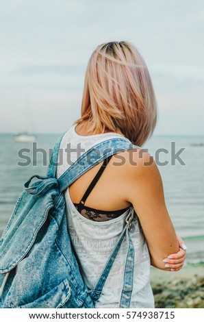 a young blonde girl walks along the riverbank , her blue jeans and denim backpack