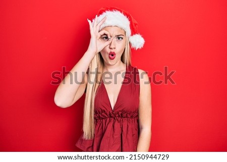 Young blonde girl wearing christmas hat doing ok gesture shocked with surprised face, eye looking through fingers. unbelieving expression. 