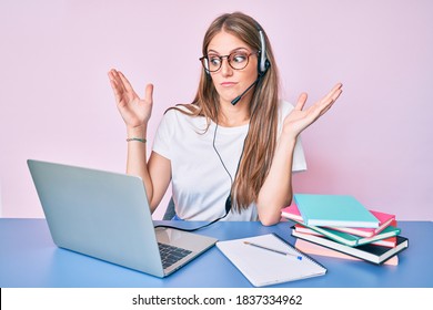 Young Blonde Girl Wearing Operator Headset At The Call Center Office Clueless And Confused With Open Arms, No Idea And Doubtful Face. 