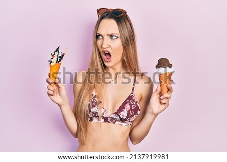 Young blonde girl wearing summer style holding ice cream angry and mad screaming frustrated and furious, shouting with anger. rage and aggressive concept. 