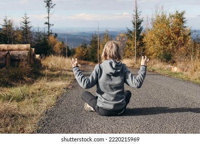 Young Blonde Hair Boy In Outdoor Clothing Sitting On Road In Yoga Position. Autumn Mountain, Hills Landscape. Fall Woodland With Yellow Birch Trees. Travel, Hiking And Reklaxation Concept. Back View