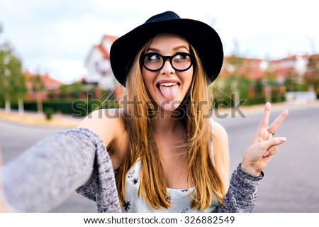 Young blonde hipster woman, making selfie, showing tongue, funny face, posing at countryside at autumn day, stylish hat and clear sunglasses.