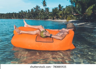 Young Blonde Man, Boy At The Beach With Lazy Inflatable Air Bed, Hammock, Couch Camping.  Lounger Sofa Beach Chair Portable Sleeping Bag Mattress. Relax, Vacation, Travel. Coconut, Sun Glasses, Hat