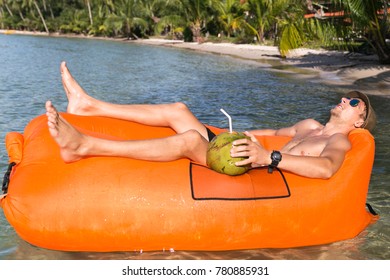 Young Blonde Man, Boy At The Beach With Lazy Inflatable Air Bed, Hammock, Couch Camping.  Lounger Sofa Beach Chair Portable Sleeping Bag Mattress. Relax, Vacation, Travel. Coconut, Sun Glasses, Hat