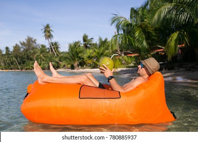 Young Blonde Man, Boy At The Beach With Lazy Inflatable Air Bed, Hammock, Couch Camping.  Lounger Sofa Beach Chair Portable Sleeping Bag Mattress. Relax, Vacation, Travel. Coconut, Sun Glasses, Hat