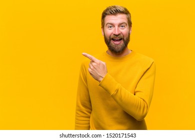 young blonde man looking excited and surprised pointing to the side and upwards to copy space against orange wall - Shutterstock ID 1520171543
