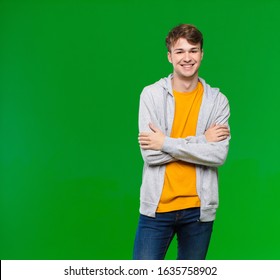 young blonde man looking like a happy, proud and satisfied achiever smiling with arms crossed against chroma key wall