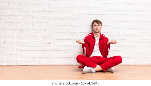 Young Blonde Man Shrugging With A Dumb, Crazy, Confused, Puzzled Expression, Feeling Annoyed And Clueless Sitting On The Floor
