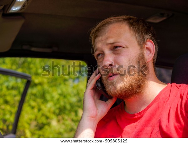 young\
blonde man speak telephone seating in his car, blonde man with\
blonde beard in red clothes speak with someone with telephone in\
right hand against car and green grass with\
shadows