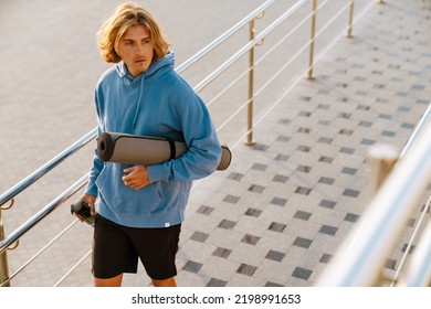 Young Blonde Man Walking With Mat And Water Bottle After Workout Outdoors