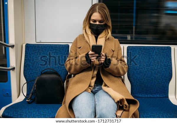 Young blonde in medical mask with phone in her hands\
sitting in subway car.