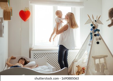 Junge blonde Mutter zweier Jungen, die in einem hellen Raum spielen. Sonniger Tag draußen. Kaukasische Familie verbringt Zeit zusammen. Älterer Bruder springt auf das Bett. Kleiner Bruder in den Händen der Mutter. Freundliche Familie