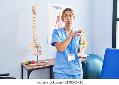 Young Blonde Orthopedic Woman Holding Anatomical Model Of Knee Joint Puffing Cheeks With Funny Face. Mouth Inflated With Air, Catching Air. 