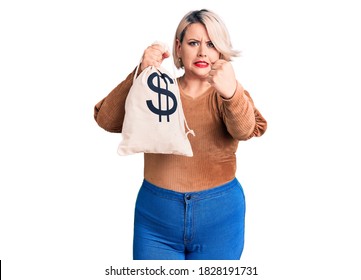 Young Blonde Plus Size Woman Holding Money Bag With Dollar Symbol Annoyed And Frustrated Shouting With Anger, Yelling Crazy With Anger And Hand Raised 