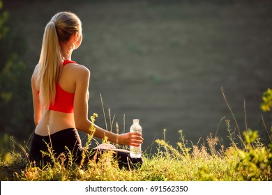 A young blonde in a red top and black pants sits on the grass in nature. A sporty woman is holding a bottle of water. Back view