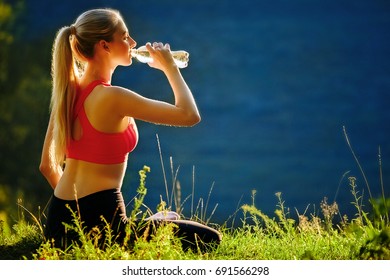 A young blonde in a red top and black pants sits on the grass in nature. A sporty woman is holding a bottle of water. Back view.Against the sea