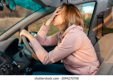 A young blonde sad woman is sitting behind the wheel of a car and holding her head in despair, covering her eyes with her hand. Side view. Stress while driving a right-hand drive car.