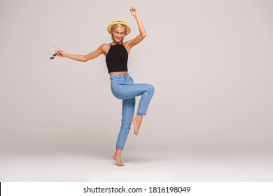 Young blonde short-haired woman wearing jeans black top and hat standing on one foot isolated on white background full body shot holding sunglasses making step laughing playful carefree copy space for