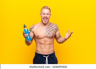 Young Blonde Strong Man With Cheerful, Carefree, Rebellious Attitude, Joking And Sticking Tongue Out, Having Fun With An Energy Drink