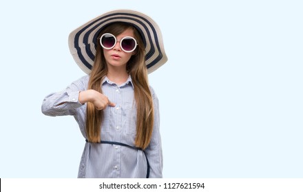 Young blonde toddler wearing hat and sunglasses with surprise face pointing finger to himself