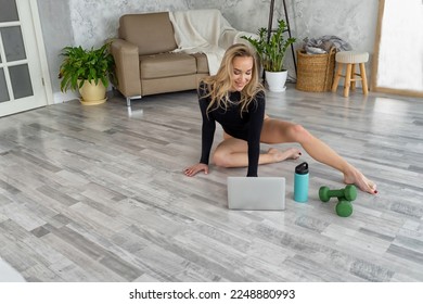 Young blonde trainer Instructor yoga using tablet teaches fitness marathon remotely distance, tutorial online. Athletic woman training stretching in black bodysuit sitting on floor with laptop at home - Shutterstock ID 2248880993