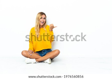 Young blonde Uruguayan woman sitting on the floor isolated on white background pointing to the side to present a product