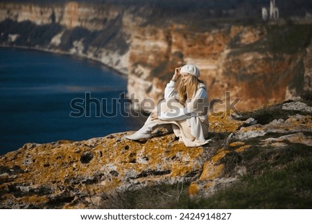 young blonde woman 35 - 40 years old in white clothes, long sweater, boots, beret sits on the ground against the backdrop of Cape Emine in Bulgaria and looks towards the sea smiling, spring, autumn