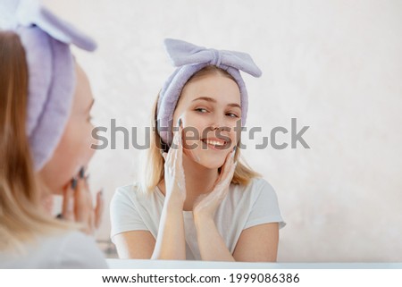 Young blonde woman applies facial Cleaner for skin wash. Teen girl washing face in morning in bathroom. Self Care morning bathroom routine woman portrait reflection in mirror.