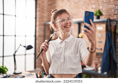 Young blonde woman artist holding paintbrush make sefie by smartphone at art studio