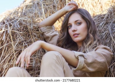 Young Blonde Woman In A Beige Jumpsuit Is Sitting On The Rolls Of Hay. Romantic Country Girl  On Hay.