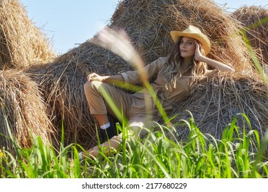 Young Blonde Woman In A Beige Jumpsuit Is Sitting On The Rolls Of Hay. Romantic Country Girl In Cowboy Hat On Hay.