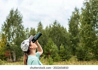 Young blonde woman bird watcher in cap and blue looking through binoculars at cloudy sky in summer forest ornithological research Birdwatching, zoology, nature, observation of animals Ornithology