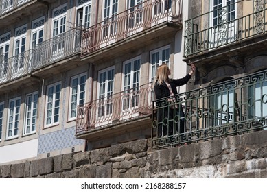 Young Blonde Woman With A Black Trench Coat Taking Selfies With Her Mobile Phone On A Balcony With Windows And Colorful Tiles Ay The Back In The City Of Porto In Portugal.