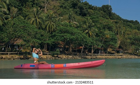 Young blonde woman in blue swimsuit rows pink plastic canoe along azure sea bay past island with palms under blue sky at resort. Traveling to tropical countries. Girl is sailing on kayak in ocean.