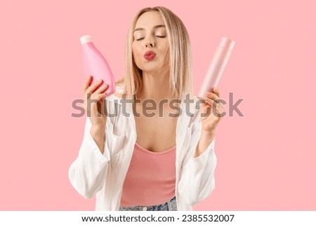 Young blonde woman with bottles of hair products blowing kiss on pink background