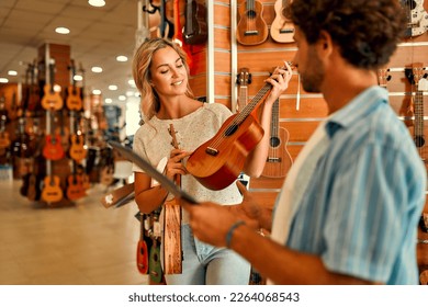 A young blonde woman buying a ukulele guitar asking for help from a sales assistant in a musical instrument store against the backdrop of walls with many different guitars. - Powered by Shutterstock