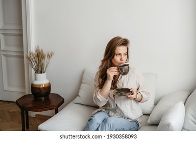 A Young Blonde Woman In Casual Comfortable Clothes Sits On A White Sofa At Home And Drinks Coffee. Scandinavian Interior, Minimalism
