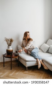 A Young Blonde Woman In Casual Comfortable Clothes Sits On A White Sofa At Home And Drinks Coffee. Scandinavian Interior, Minimalism