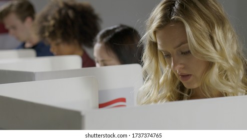 Young Blonde Woman Considers Her Vote In Booth With Others At Polling Station.  US Flag On Wall In Background.