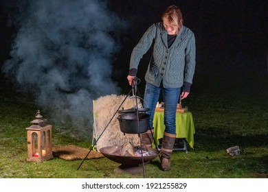 Young blonde woman is cooking dinner in a black kettle, outside on a winter evening. She is camping in the forest in a cozy setting, Night colorful landscape.