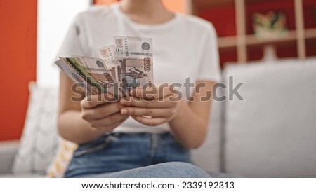 Young blonde woman counting russia rubles banknotes sitting on sofa at home